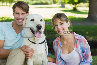 Happy couple sitting with their labrador in the park smiling at camera