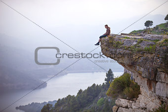 Young man sitting on edge of cliff and looking at river below
