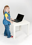 little girl in blue jeans standing at a table with a laptop