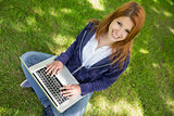 Pretty redhead smiling at camera in the park using laptop