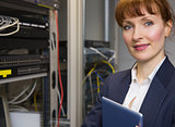 Pretty technician smiling at camera beside open server holding tablet pc