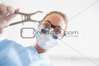 Dentist in surgical mask and dental loupes holding pliers over patient