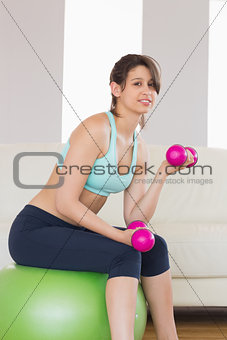 Fit brunette sitting on exercise ball lifting hand weights