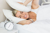 Woman covering ears with pillow and alarm clock in foreground