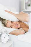 Woman covering ears with pillow and alarm clock in foreground