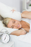 Woman covering ears with pillow and alarm clock on table