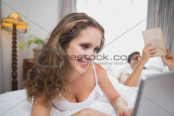 Happy woman using laptop in bed with man in background