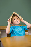 Happy pupil holding book on his head at desk