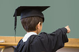 Cute pupil in graduation robe pointing in classroom