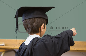Cute pupil in graduation robe pointing in classroom