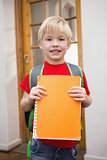 Cute pupil smiling at camera in classroom holding notepad