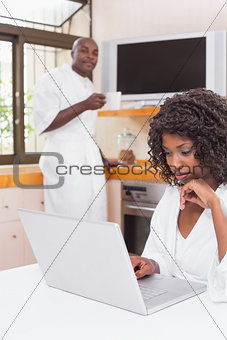 Pretty woman in bathrobe using laptop at table with partner in background