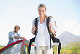 Attractive hiking blonde smiling at camera while partner pitches tent