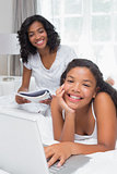 Mother reading magazine with daughter using laptop on bed