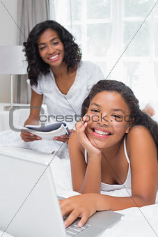 Mother reading magazine with daughter using laptop on bed