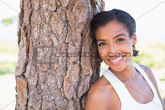 Fit woman leaning against tree smiling at camera