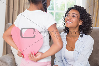 Pretty mother sitting on couch with daughter hiding heart card