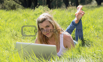 Pretty blonde lying on grass using laptop smiling at camera