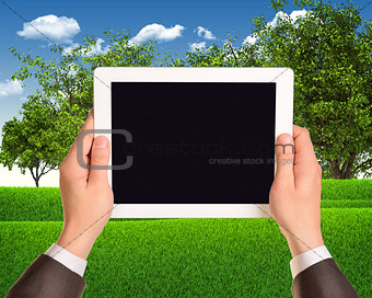 Digital tablet in hands with grass field and trees as backdrop