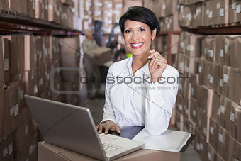 Pretty warehouse manager smiling at camera using laptop