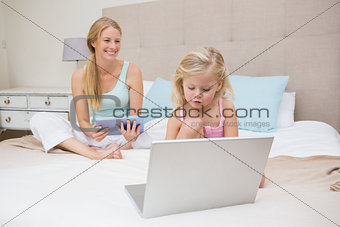 Cute little girl and mother on bed using tablet and laptop