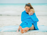 Smiling mother and baby girl wrapped in towel sitting on beach