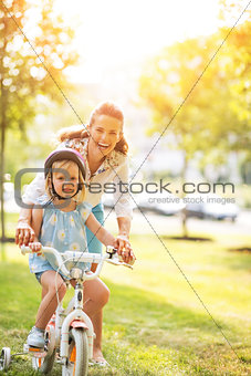 Portrait of happy mother helping baby girl riding bicycle in par