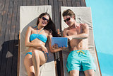 Couple using digital tablet on sun loungers by swimming pool