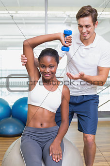 Personal trainer helping client lift dumbbell on exercise ball