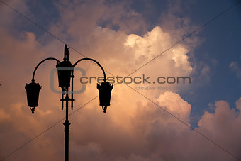 Silhouette of black street lamp against background bright multi colored sunset clouds in the sky.