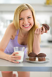 Happy teenager girl eating muffin with milk in kitchen