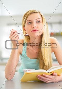 Young woman eating yogurt in kitchen and reading book