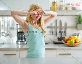 Young woman standing in in kitchen and rubbing eyes after sleep