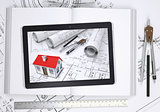Small house with drawings displayed on tablet screen. Open book and tools of architect
