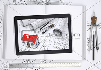 Small house with drawings displayed on tablet screen. Open book and tools of architect