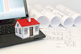 Small model house with red roof on laptop near scrolls of architectural drawings