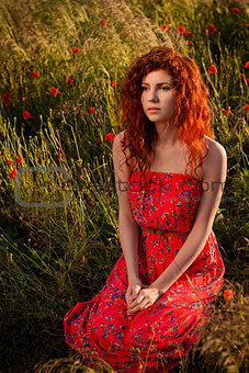 Red-haired girl sitting in the poppies meadow at sunset