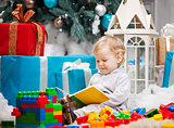 Cute toddler boy sitting at Christmas tree and reading book. Building blocks scattered around.
