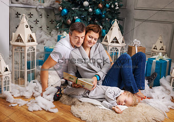 Couple reading book to toddler son beside Christmas tree, boy falling asleep