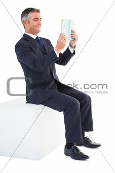Smiling businessman sitting on a cube reading a book