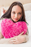 Pretty brunette holding heart cushion on bed