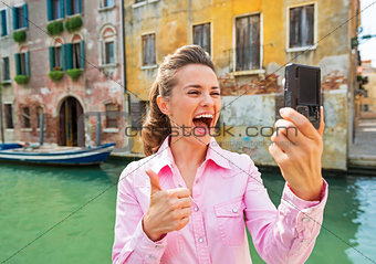 Happy young woman showing thumbs up and making selfie in venice,