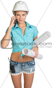 Beautiful girl in white helmet, shorts with shirt holding scrolls drawings and talking on phone
