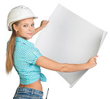 Beautiful woman builder looking at camera and holding in front of him large paper sheet. Rear view