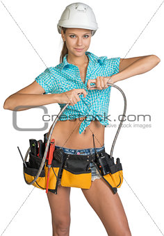Pretty girl in helmet, shorts, shirt and tool belt with tools connects two flexible hose for plumbing
