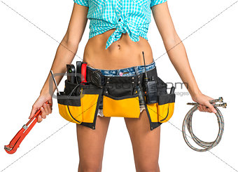 Plumber in shorts, shirt, tool belt with tools holding flexible hose and wrench