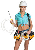 Pretty electrician in helmet, shorts, shirt, tool belt with tools holding screwdriver and an electric cable