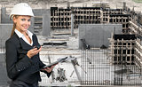 Beautiful businesswoman in helmet holding paper holder and portable radio. Construction site as backdrop