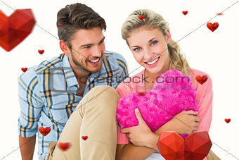 Composite image of attractive young couple sitting holding heart cushion