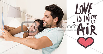 Composite image of attractive couple lying in bed with tablet pc